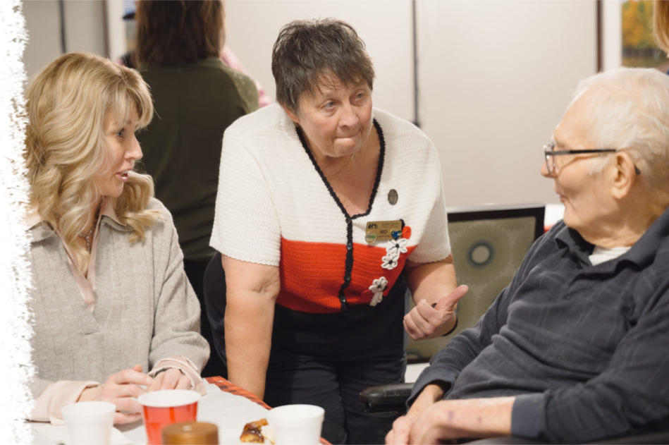 Three people engage in conversation around a table with cups, as one person leans in to speak with another.
