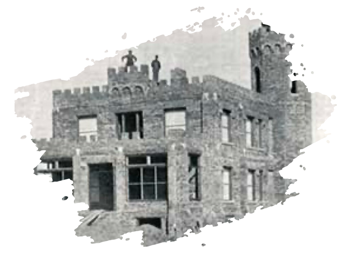 A black-and-white photo of a stone, castle-style building with two figures standing on its roof.