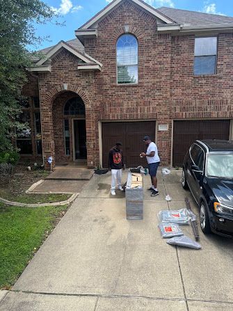 People unloading boxes and supplies in a driveway beside a brick house with an open garage.