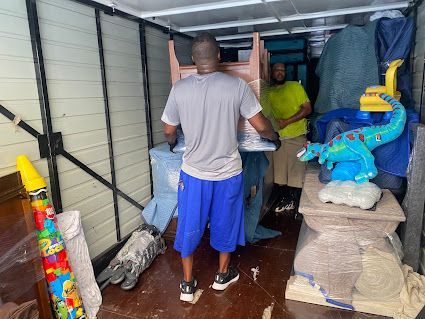 People packing furniture and bags into a storage unit, with stacked household items and toys nearby