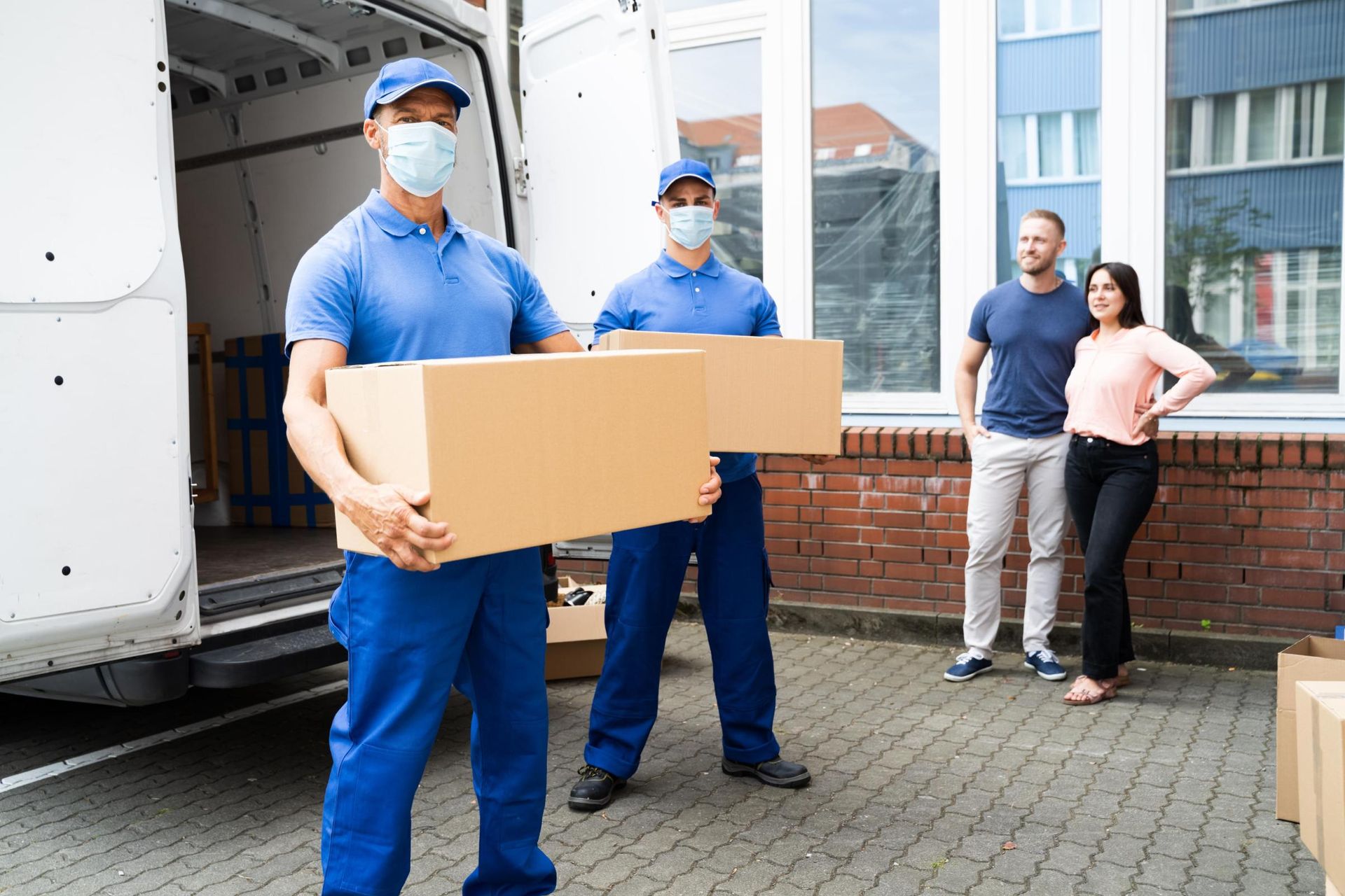 Two masked movers in blue uniforms carry a cardboard box outside a building.