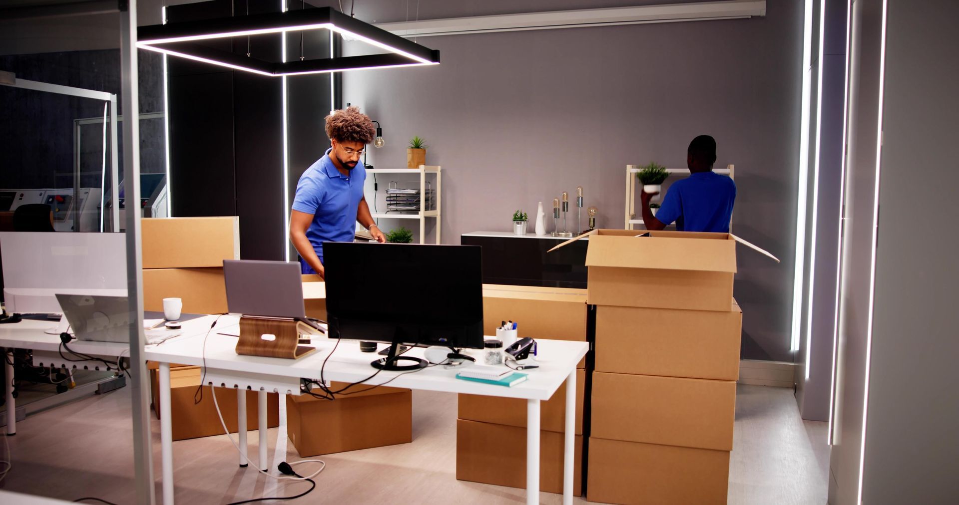Two people packing boxes in a modern office with desks, monitors, and a hanging light fixture.