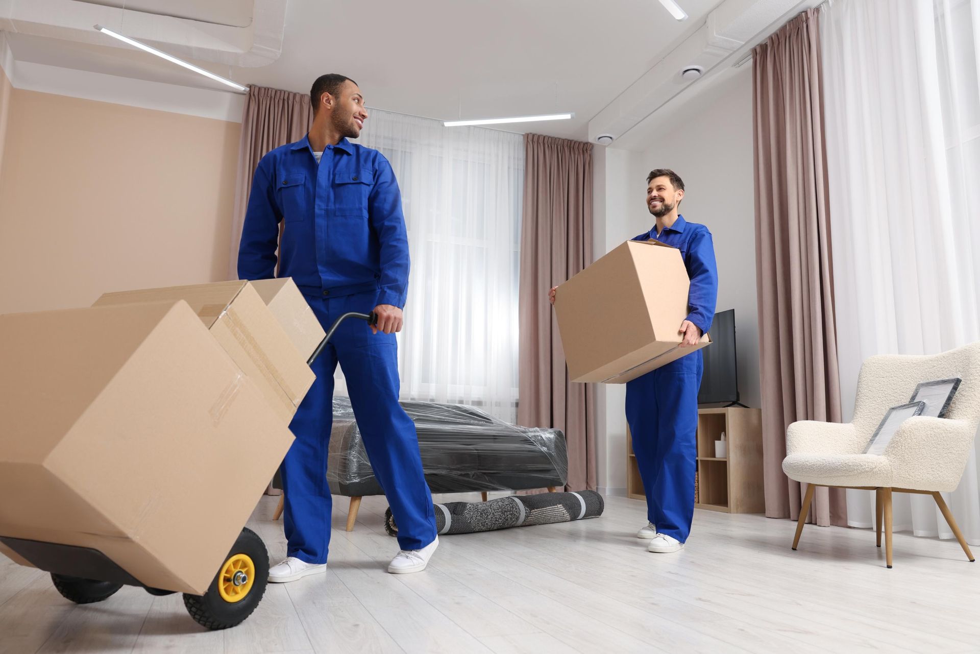 Two movers in blue uniforms carrying cardboard boxes in a bright living room