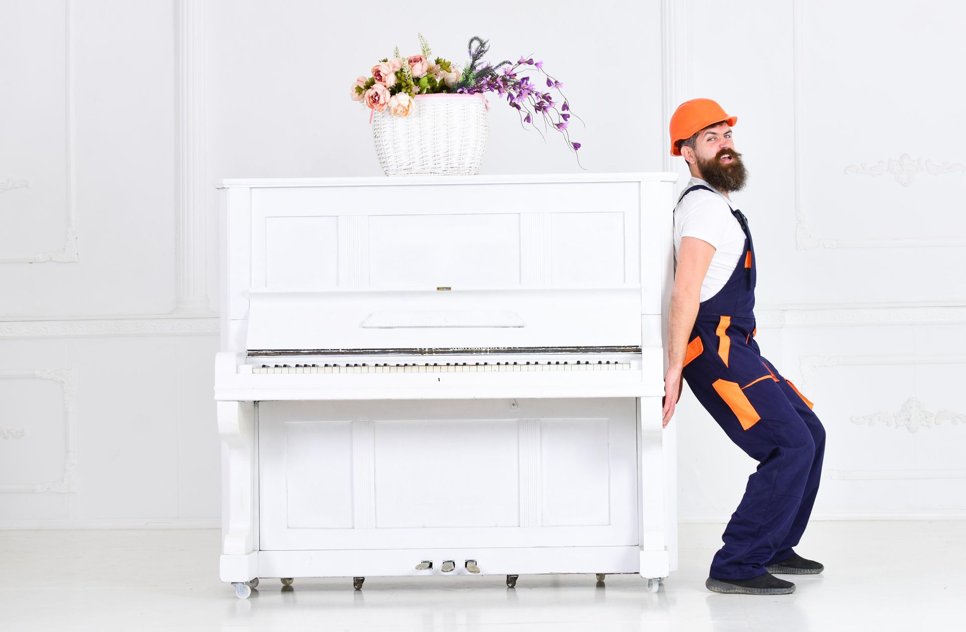 White piano with flowers; construction worker in orange hard hat leans back beside it in a bright white room.