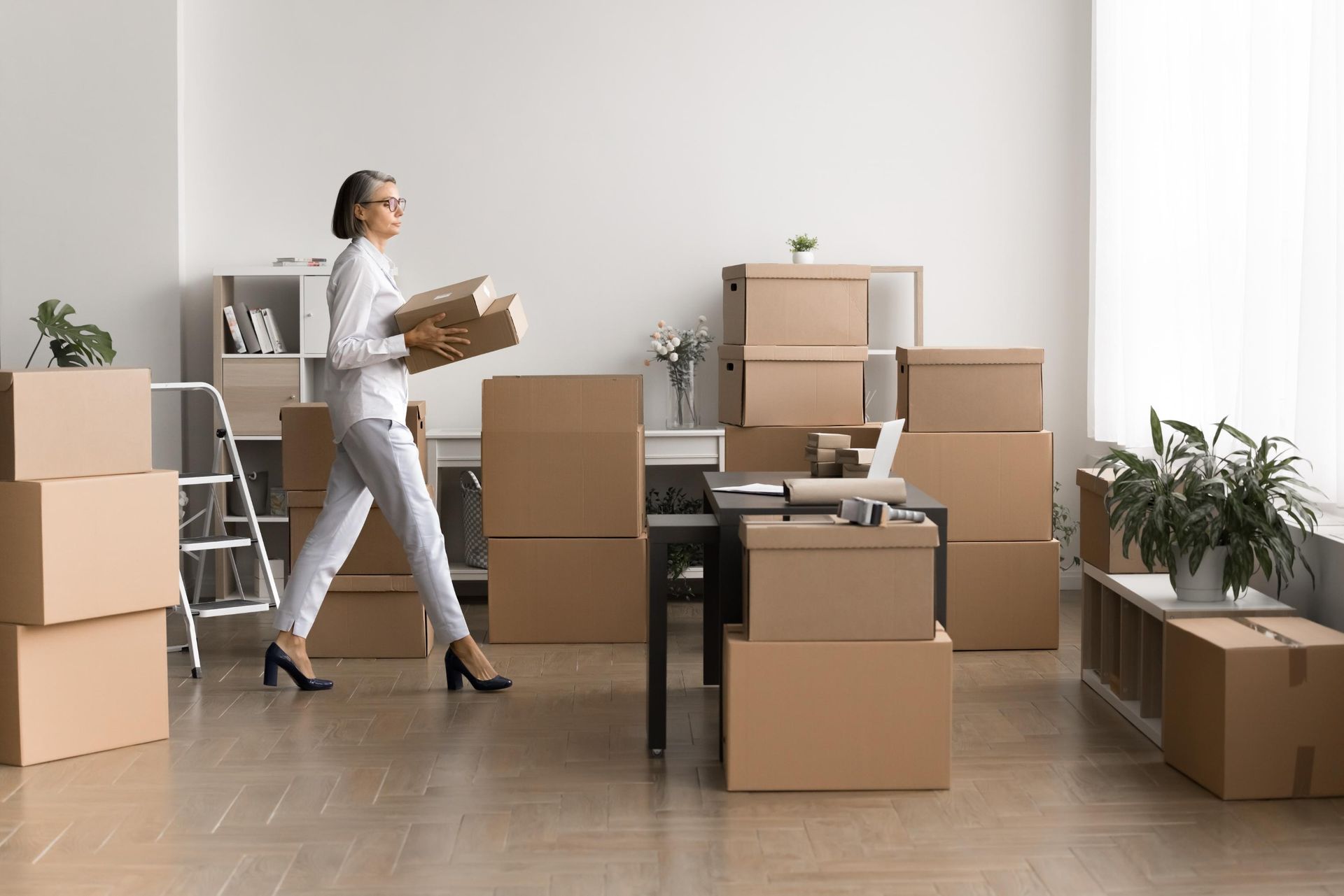Woman carrying boxes in a nearly empty room with moving boxes and plants