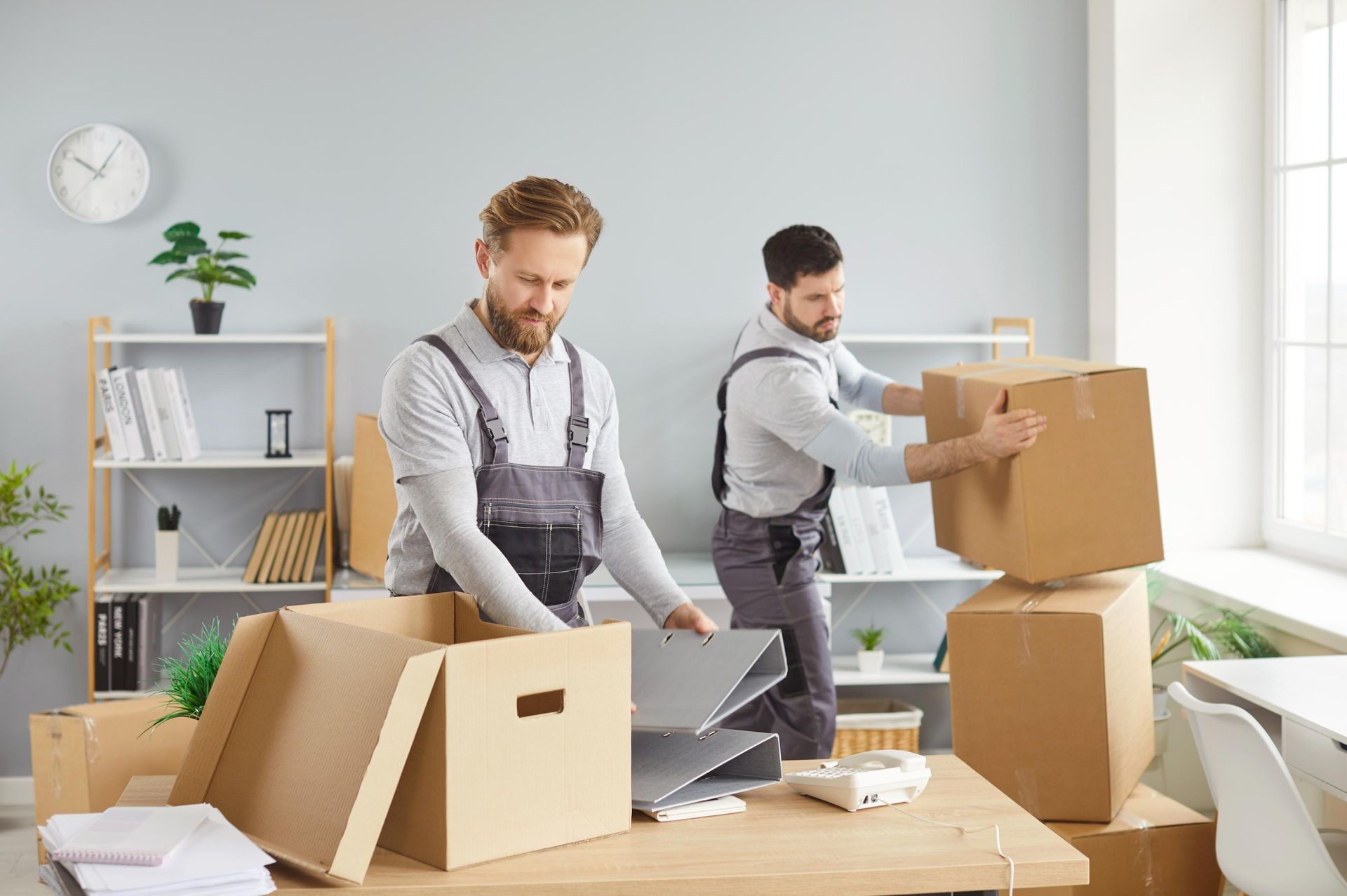 Two men packing boxes in a bright office with laptops and shelves nearby.
