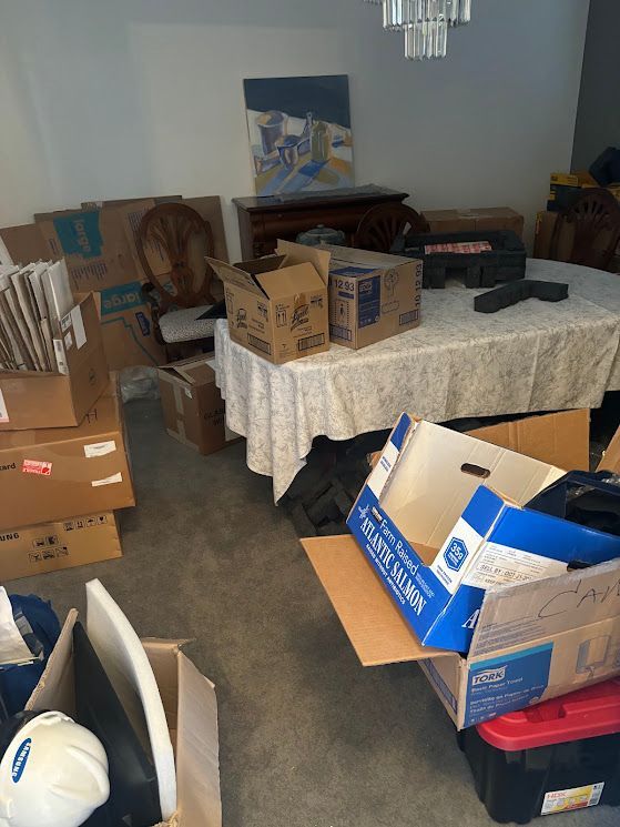 Cluttered room with stacked moving boxes, a table covered in a white cloth, and assorted packed items.