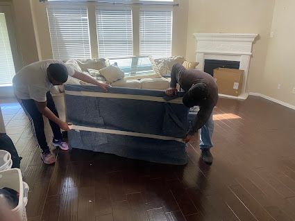 Two people moving a large couch in a living room with dark wood floors and a fireplace