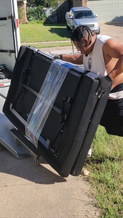 Person in a helmet carrying a large black folding table wrapped with straps on a driveway.