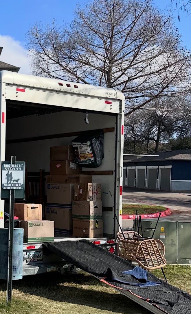 Moving truck being unloaded with boxes on a ramp in a driveway, trees and houses in the background