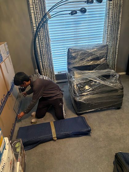 Person kneeling beside a rolled blue sleeping mat in a room with wrapped furniture and a window blind.
