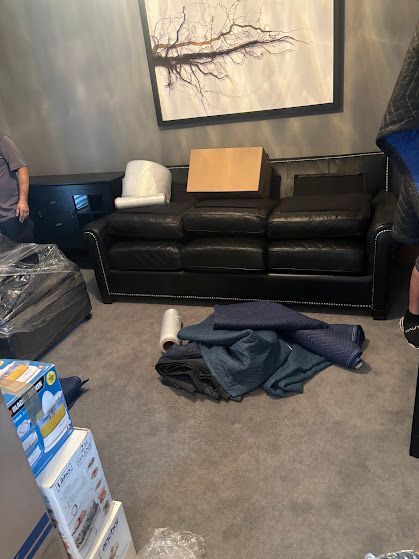 Black leather couch in a room with scattered clothes, boxes, and a painting on the wall.