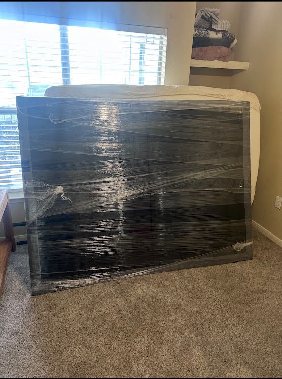 Large black item wrapped in plastic leaning against a wall in a carpeted room near a window.