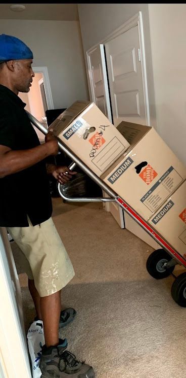 Man in blue cap pushes hand truck stacked with moving boxes through a hallway