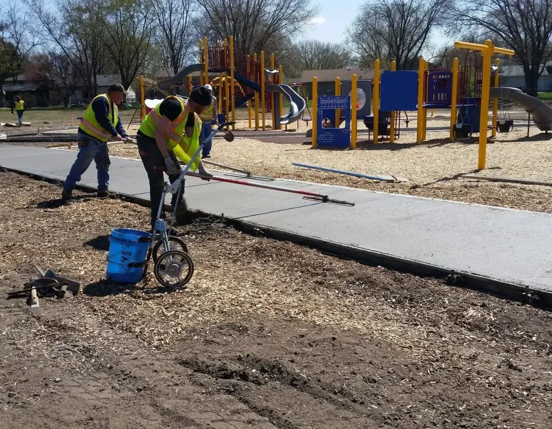 A group of construction workers are working on a concrete walkway in front of a playground.