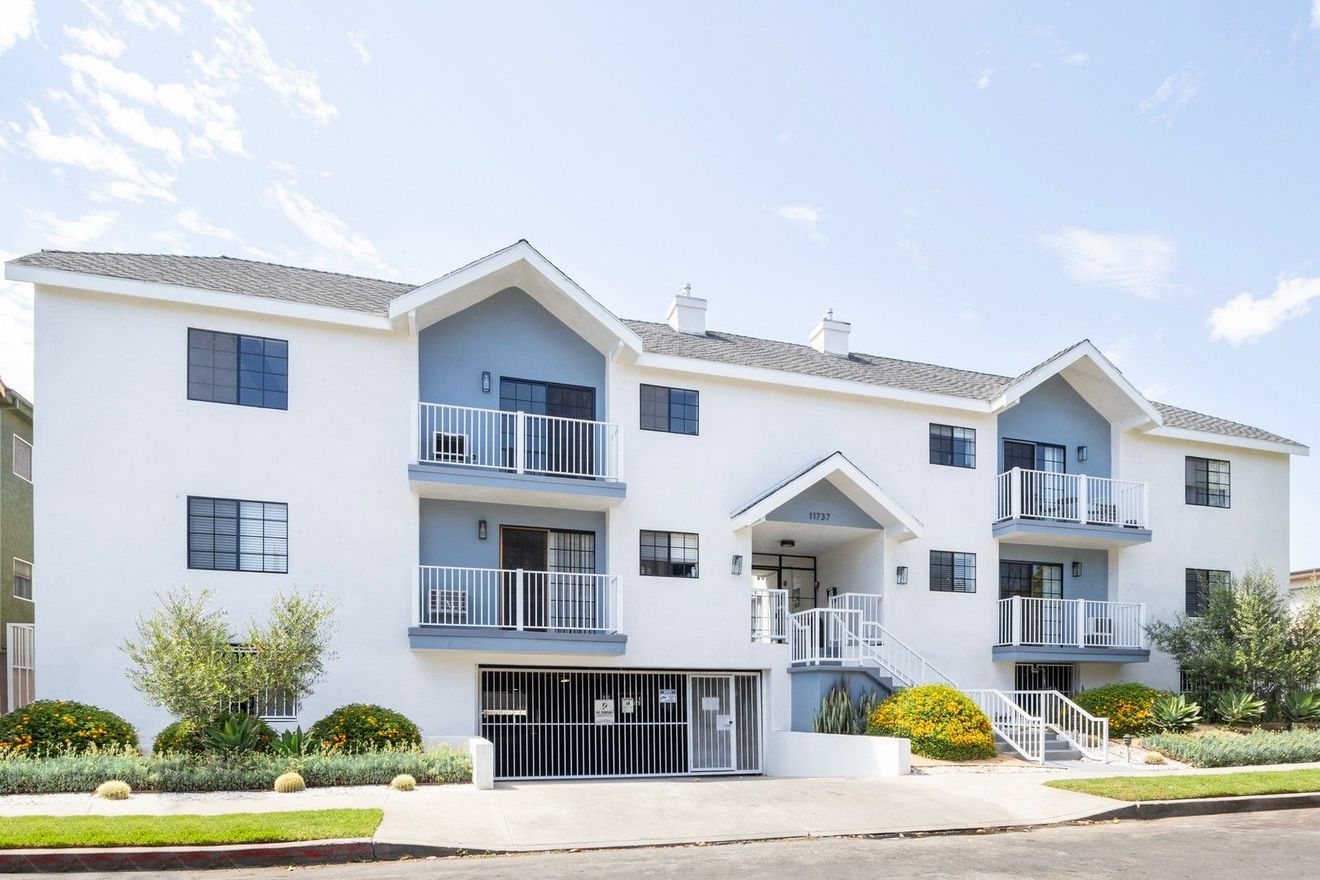 Apartment building with balconies and beige stucco exterior, viewed from a sunny landscaped courtyard