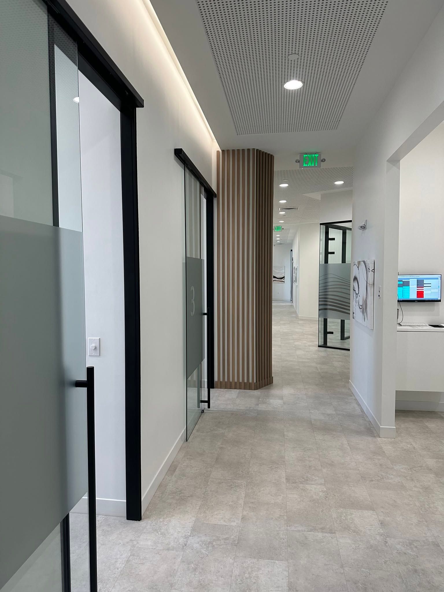 Modern office hallway with glass-walled rooms, beige carpet, patterned ceiling, and green exit sign.
