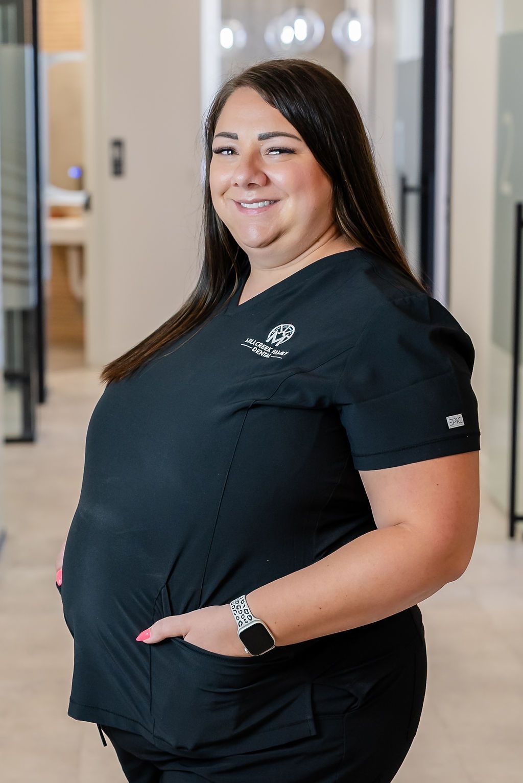 Pregnant person in black scrubs standing indoors and smiling, with hands on belly