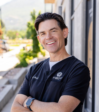 Smiling person in navy scrubs standing outdoors with arms crossed near a building.