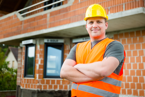 Construction worker in yellow helmet and orange vest, arms crossed, in front of a brick building.
