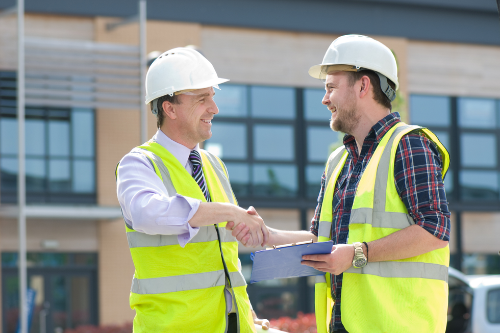 Two men in hard hats and vests shake hands on a construction site.