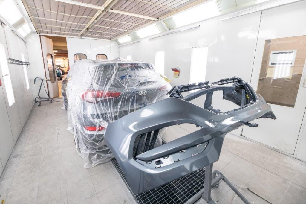 A car wrapped in plastic inside a paint booth; detached grey bumper in foreground.