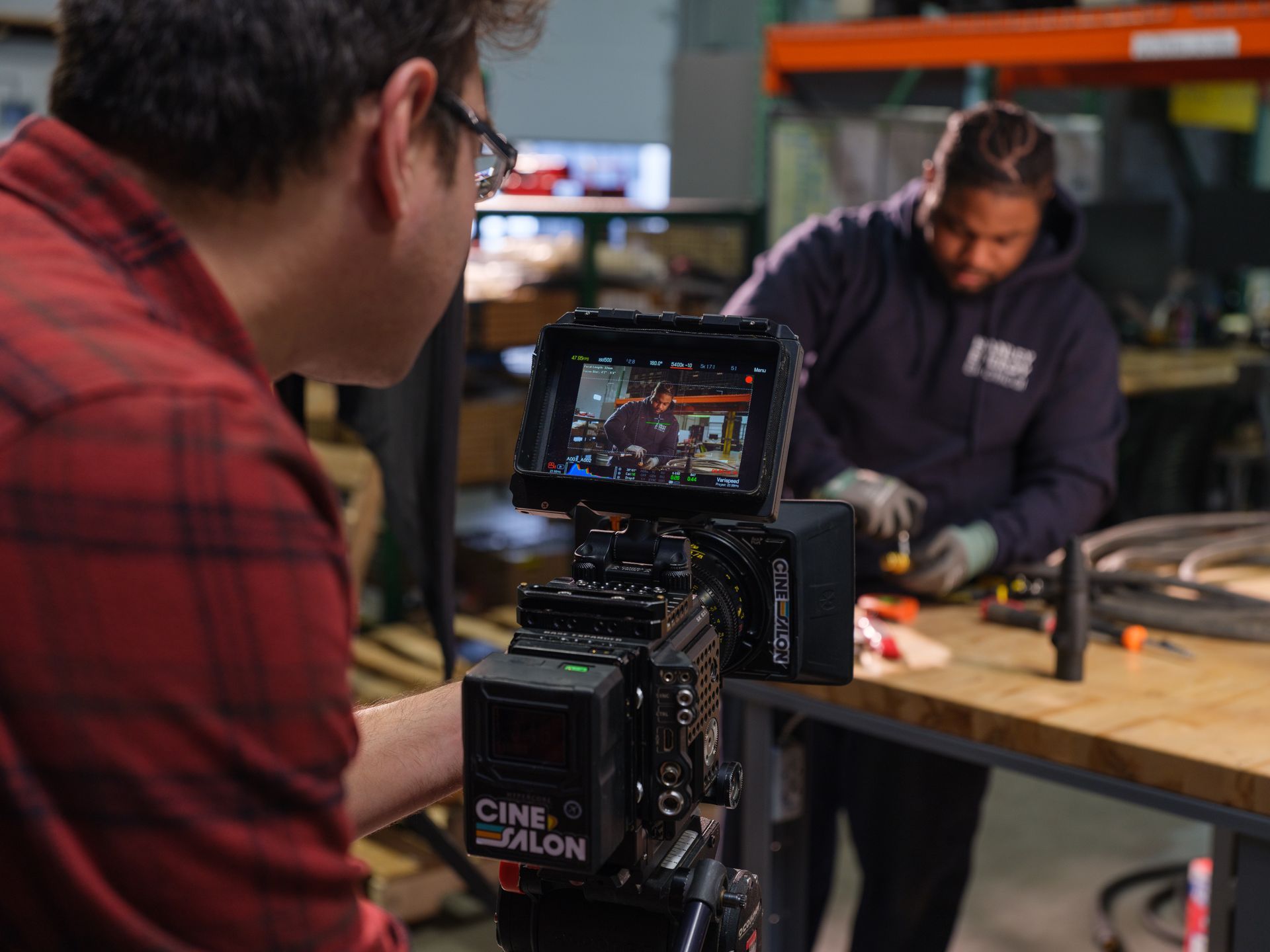 A person filming a person working in a workshop. The camera displays the scene.