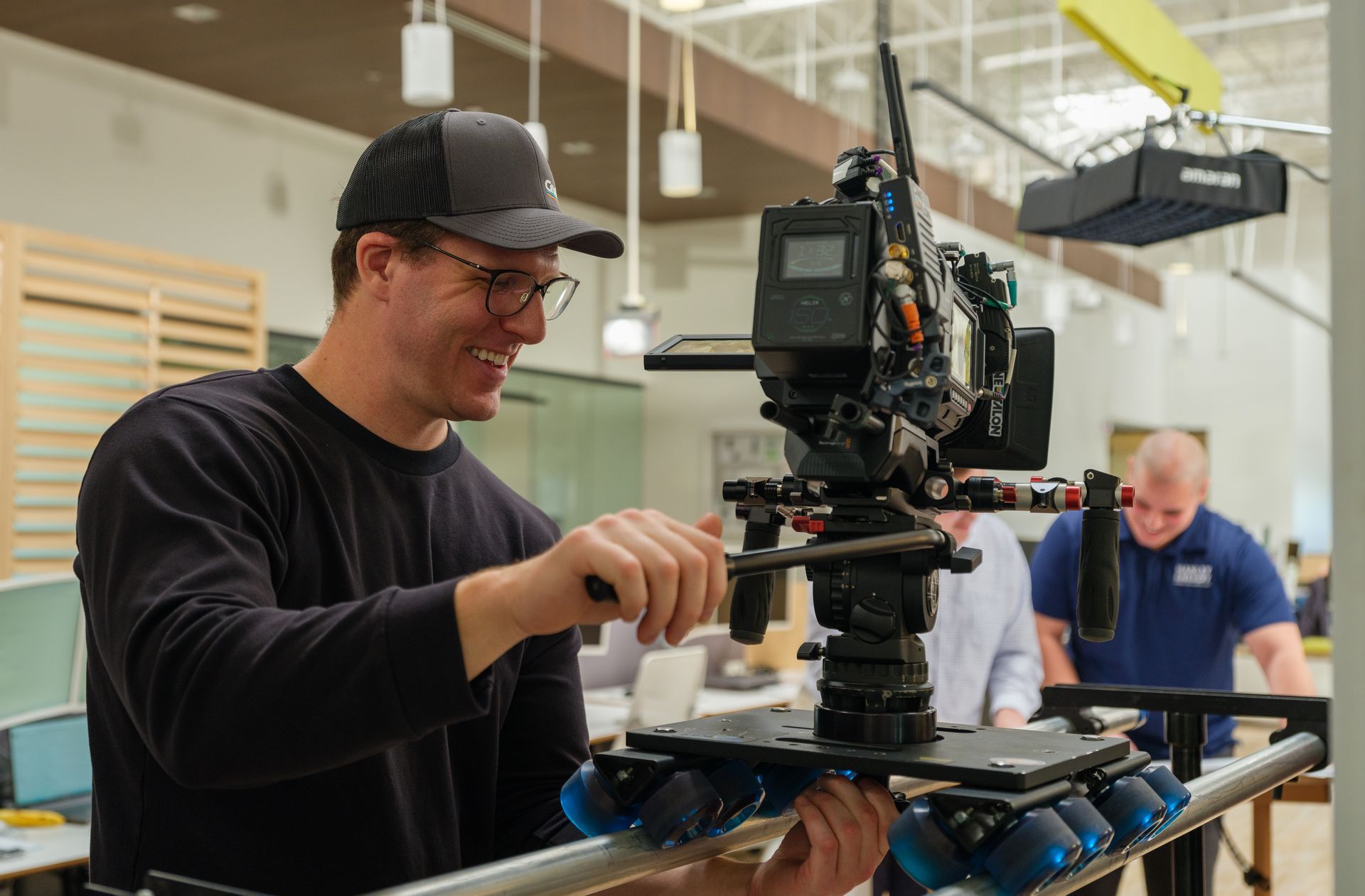 Person adjusting a large video camera on a sliding track; another person in background.
