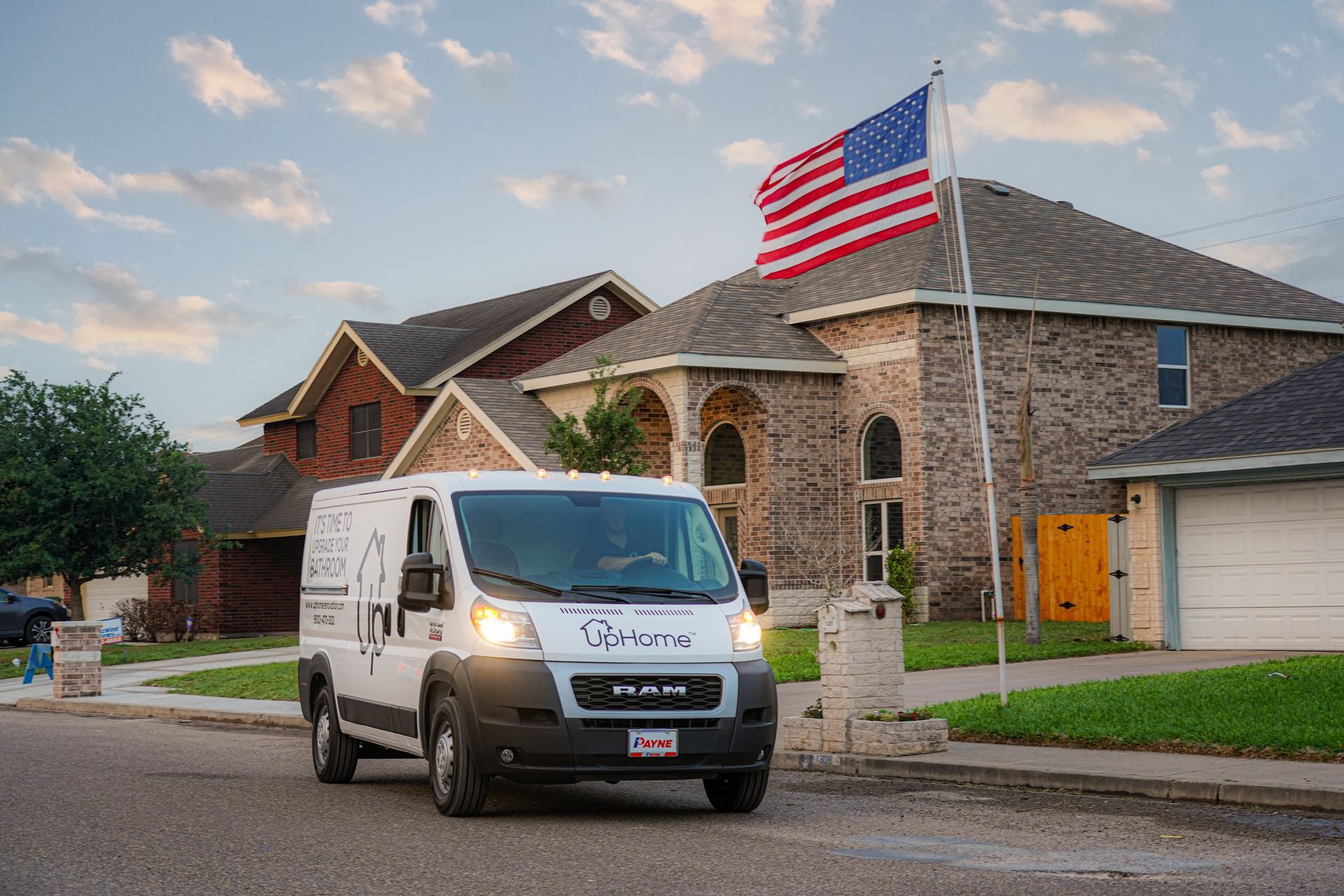 A white van is driving down a street in front of a house with an american flag on the roof.
