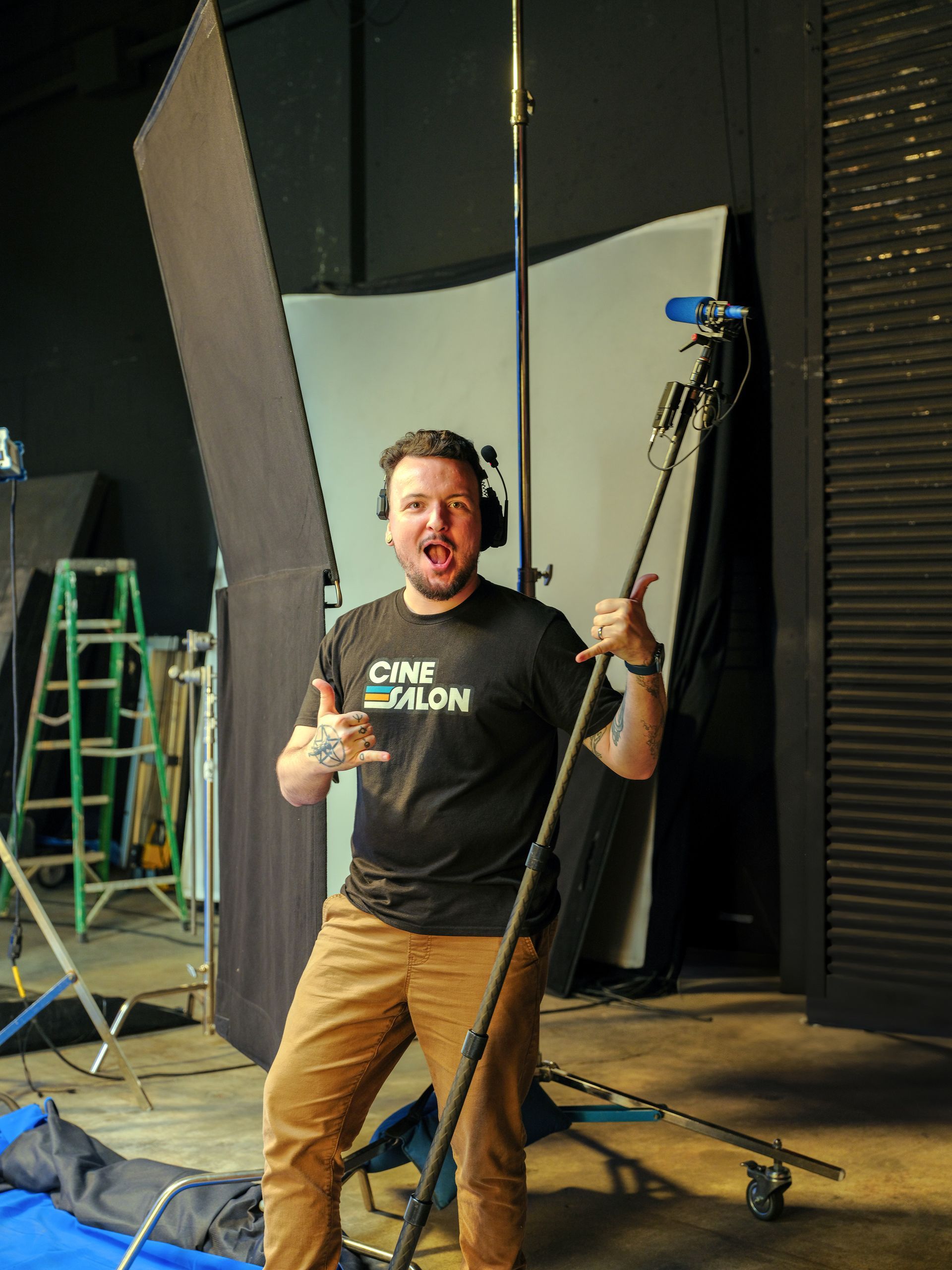 Man holding a boom mic in a studio, giving a thumbs-up. He wears a black CineSalon shirt, brown pants. Background: lights, equipment.
