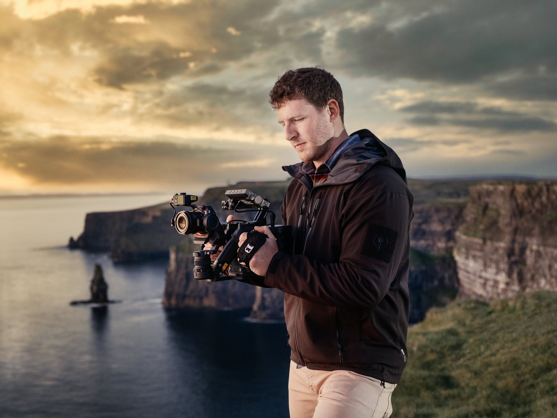 Shane Yeager filming with a camera on a cliff overlooking ocean in Ireland. Cloudy sky, rocks in the background.