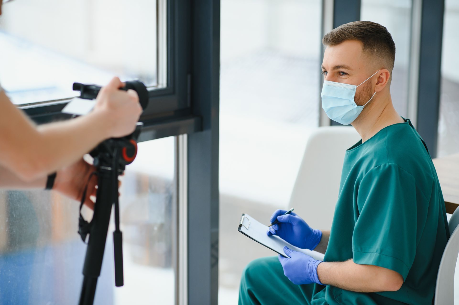 A doctor wearing a mask and gloves is sitting in front of a camera.