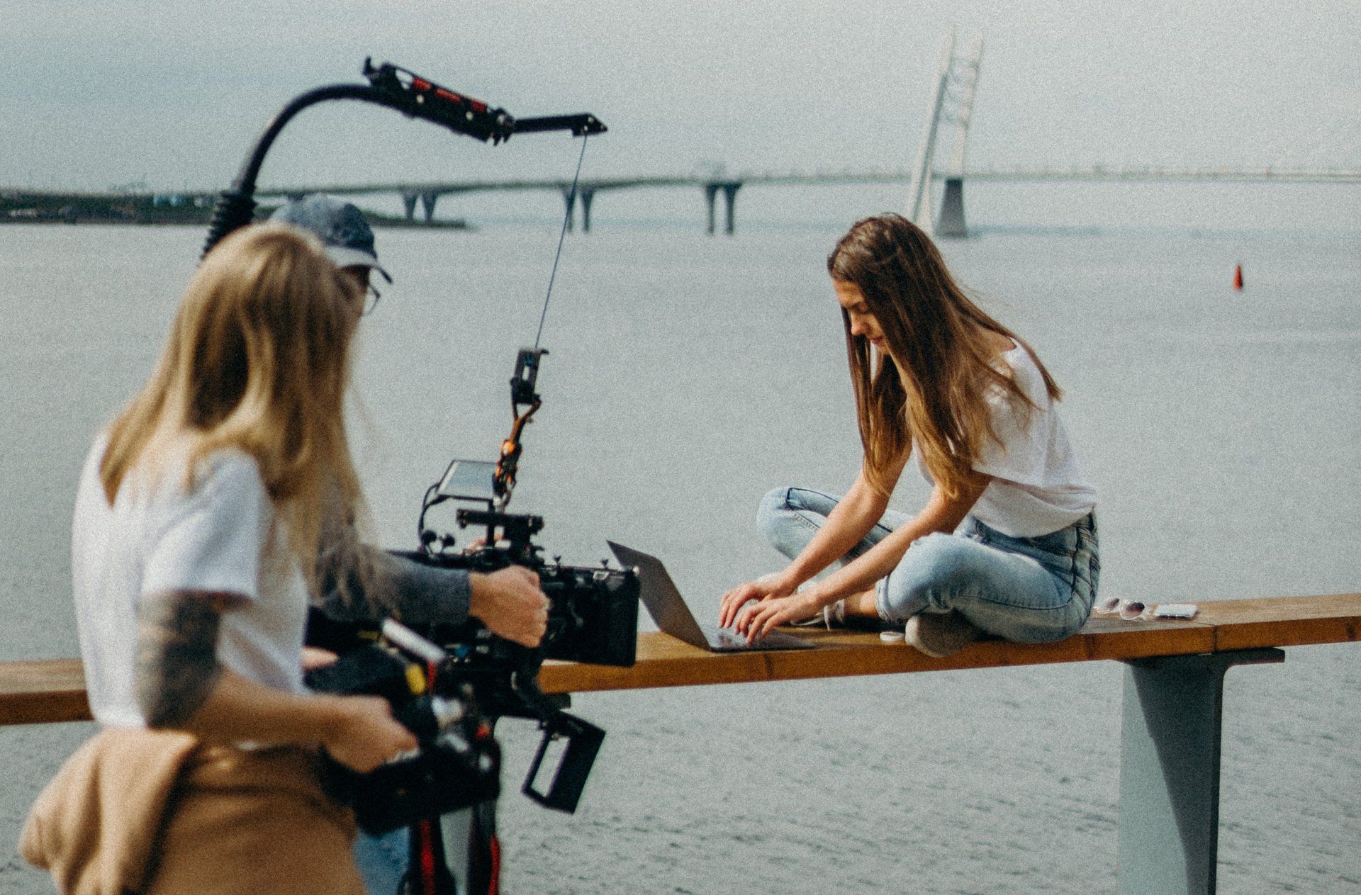A woman is sitting on a bench with a laptop and a cameraman is recording her.
