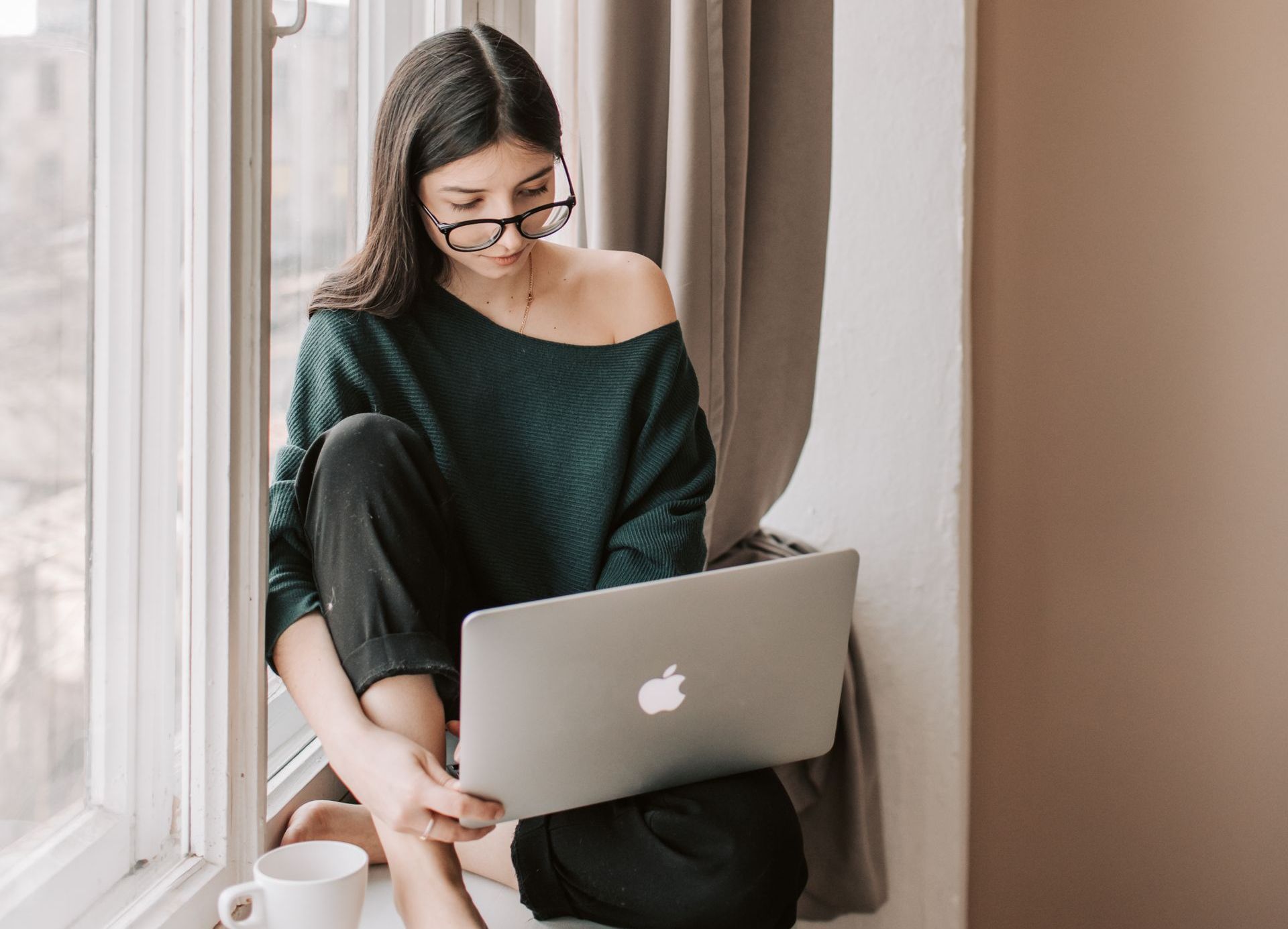 A woman is sitting on a window sill using a laptop computer.