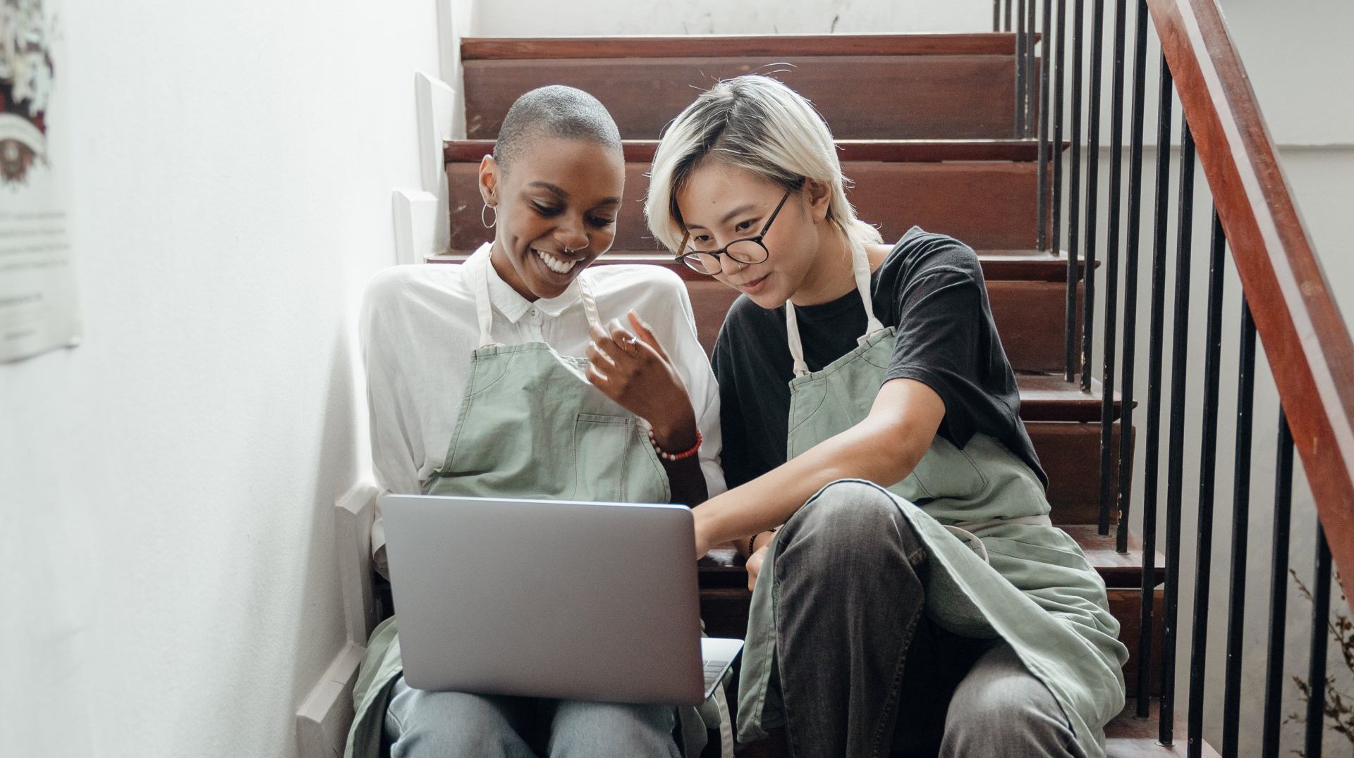 Two women are sitting on the stairs looking at a laptop.
