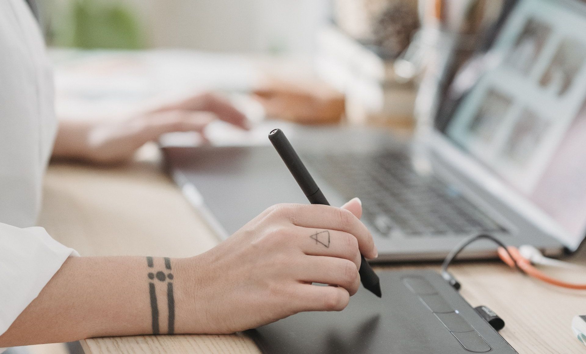 A woman is using a pen on a tablet in front of a laptop computer.