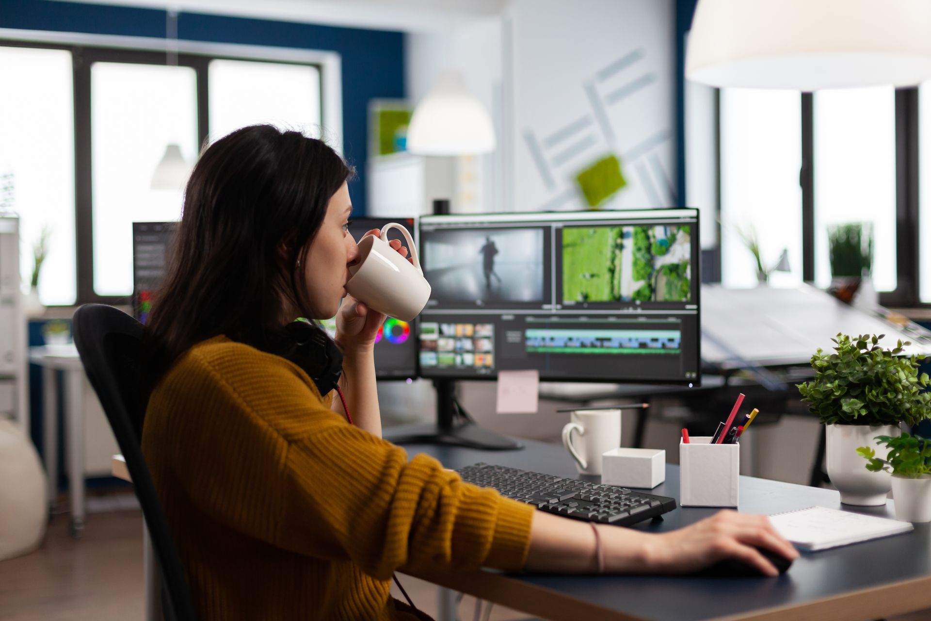 A woman is drinking coffee while sitting at a desk in front of a computer.