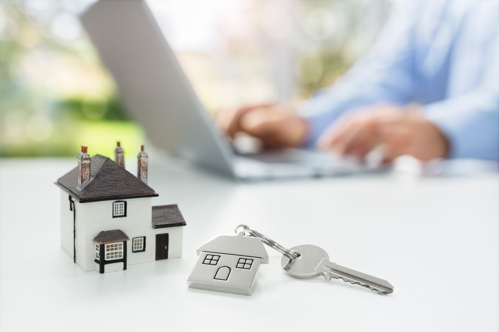 A man is typing on a laptop next to a model house and keys.