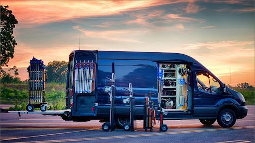 Dark blue van with open back, revealing equipment. Carts with stands and equipment are beside it. Sunset in the background.