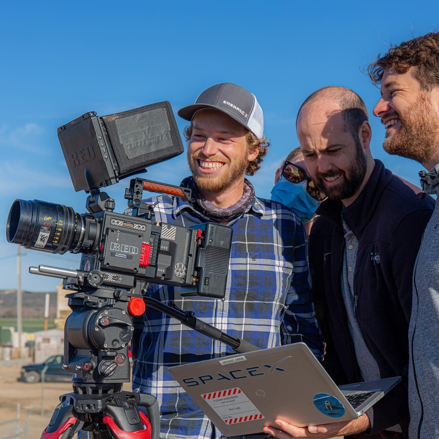 Three men smile, looking at a professional video camera on a tripod outdoors, one holding a laptop.