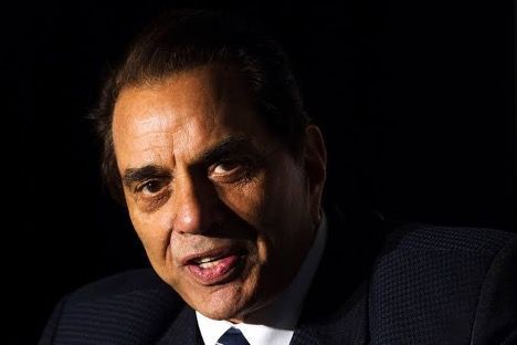 Man in a dark suit speaking, lit by strong overhead light.