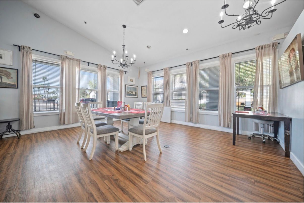 Bright dining room with a white table and chairs, large windows, chandeliers, and wood flooring