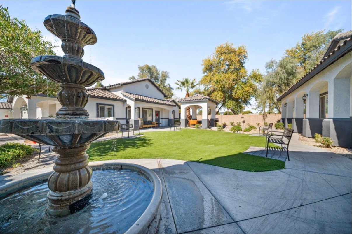 Courtyard fountain beside white stucco buildings, green lawn, and patio chairs under a blue sky