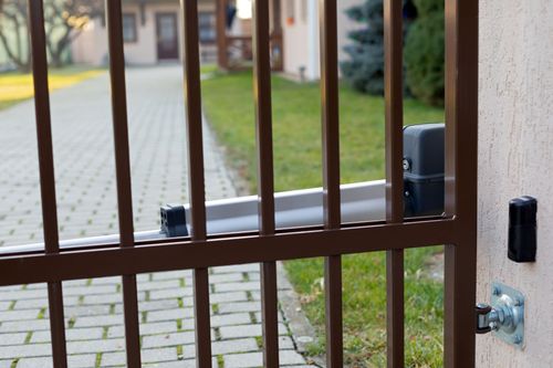 Brown metal gate with vertical bars, view of a paved walkway and courtyard beyond, with a door lock on the right