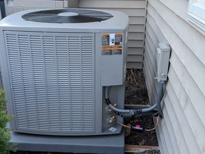 Outdoor central air conditioner unit beside a house with visible wiring and pipes near the foundation