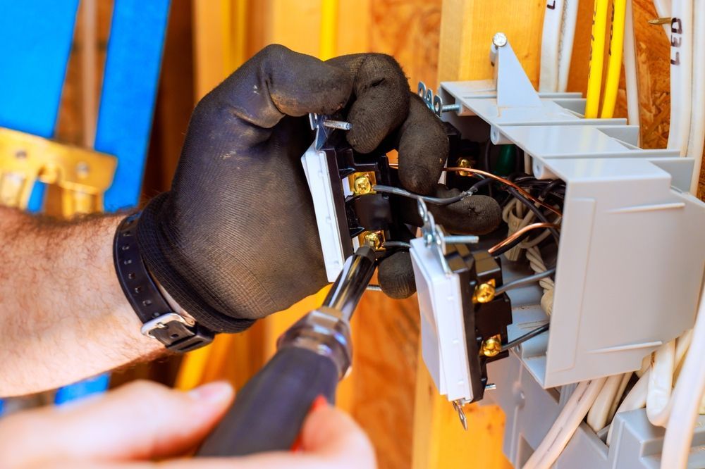 Gloved hands using a screwdriver to repair an electrical panel mounted on a wooden wall