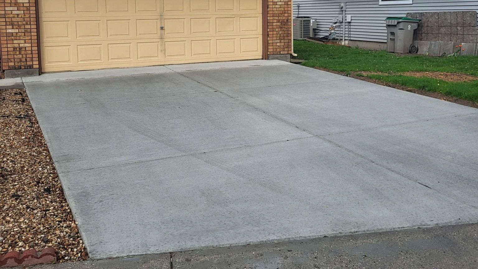 Freshly poured concrete driveway in front of a beige garage, bordered by gravel landscaping and a gr