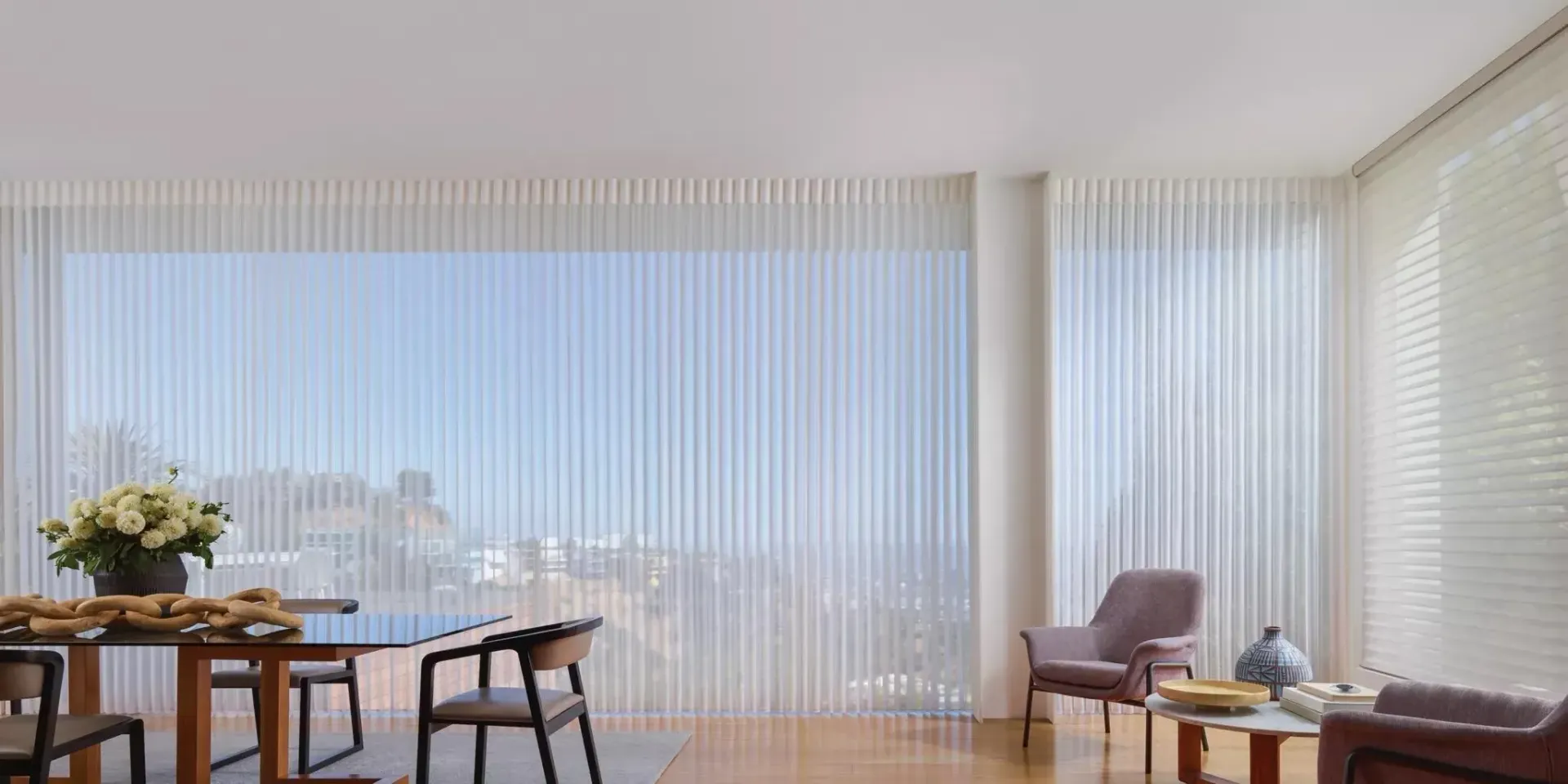A modern dining room with large windows, sheer white curtains, and a wooden dining table.