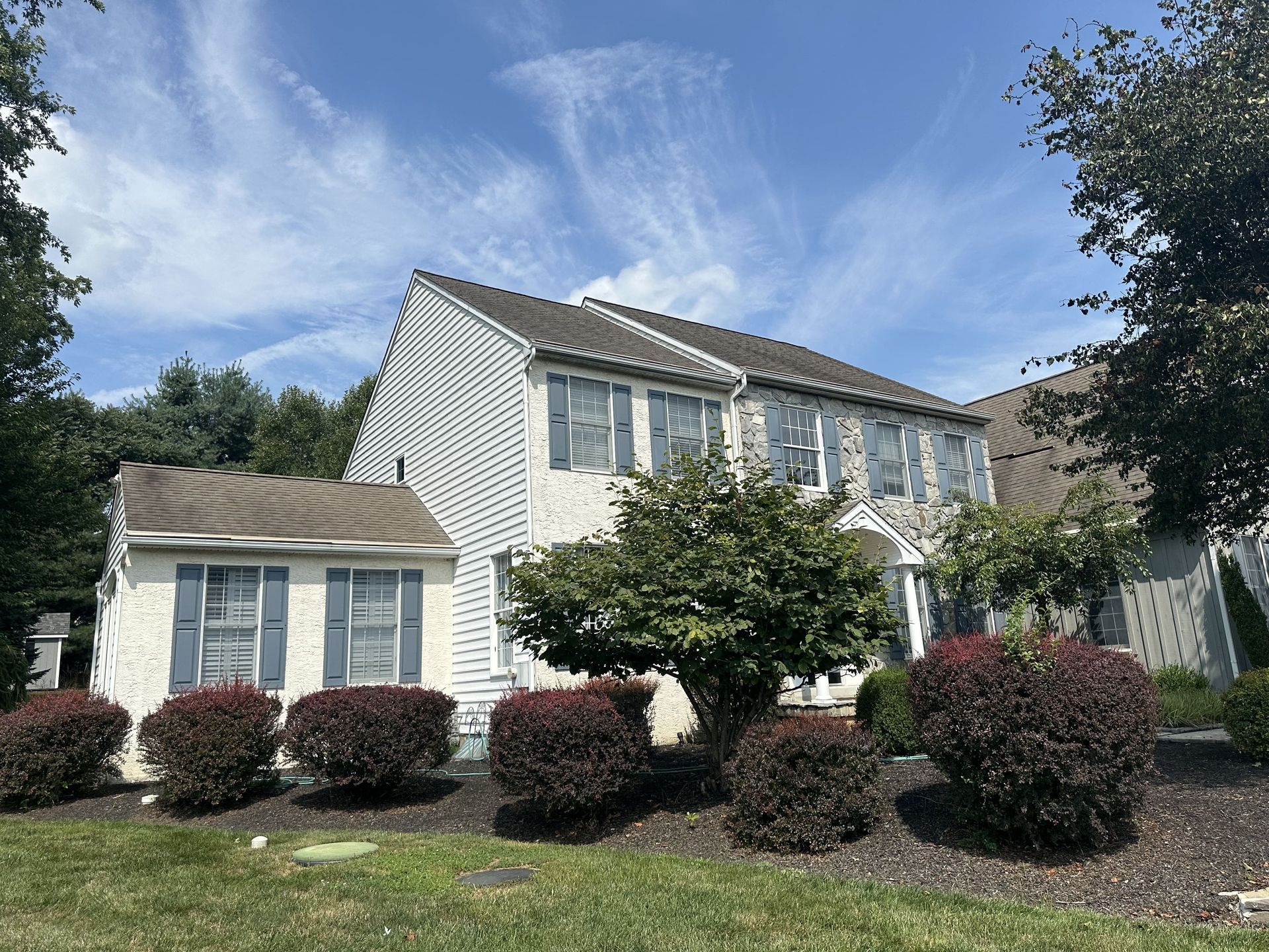 A white house with a brown roof is surrounded by bushes and trees.