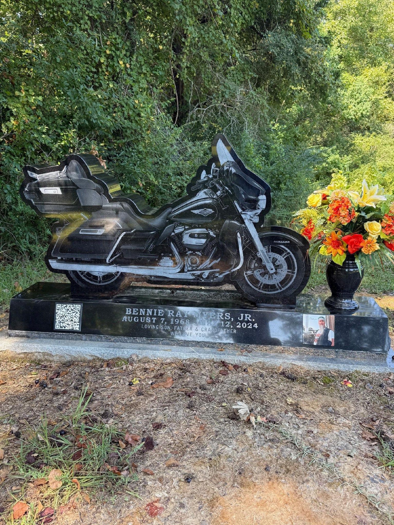 A black granite tombstone shaped like a touring motorcycle, decorated with colorful flowers, set in a wooded cemetery.